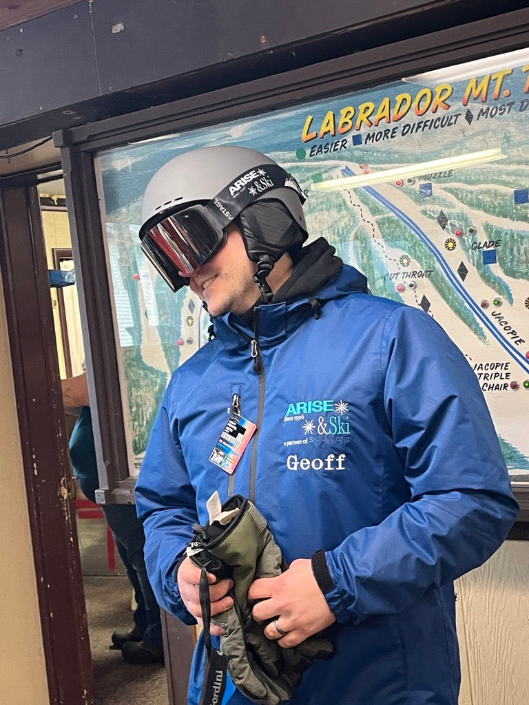 A man wearing a blue ARISE & Ski volunteer jacket, a grey ski helmet, and a pair of STAGE Custom Ski Goggles. The ski goggles feature ARISE & Ski's logo on the strap.