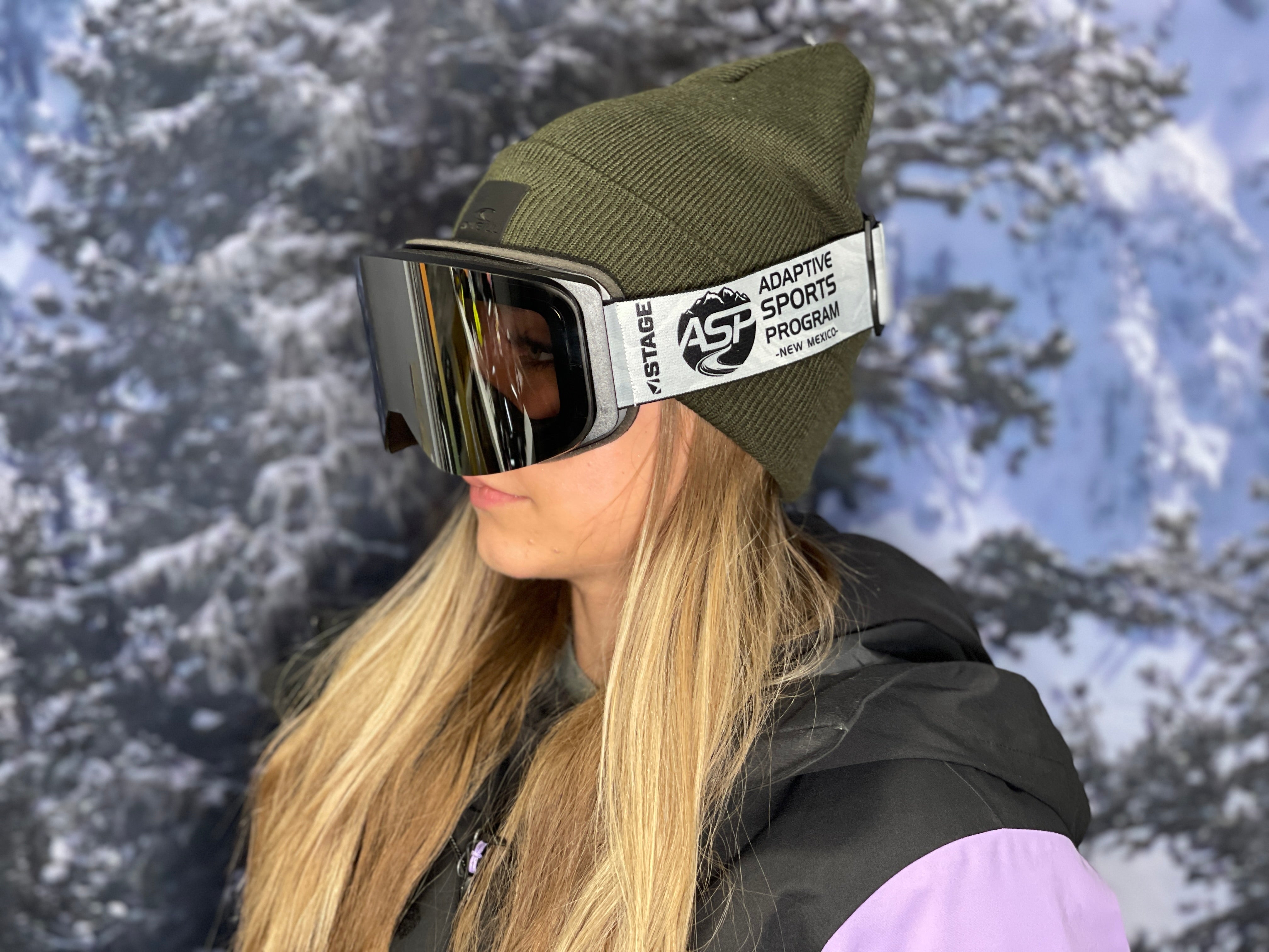 A woman wearing a pink and black ski jacket, green beanie, and STAGE custom ski goggles, standing against a snowy backdrop. The goggles feature "ASP Adaptive Sports Program - New Mexico" logo on the strap and a Mirror Chrome Smoke lens. The model of the goggle is the STAGE Frameless Prop Ski Goggle