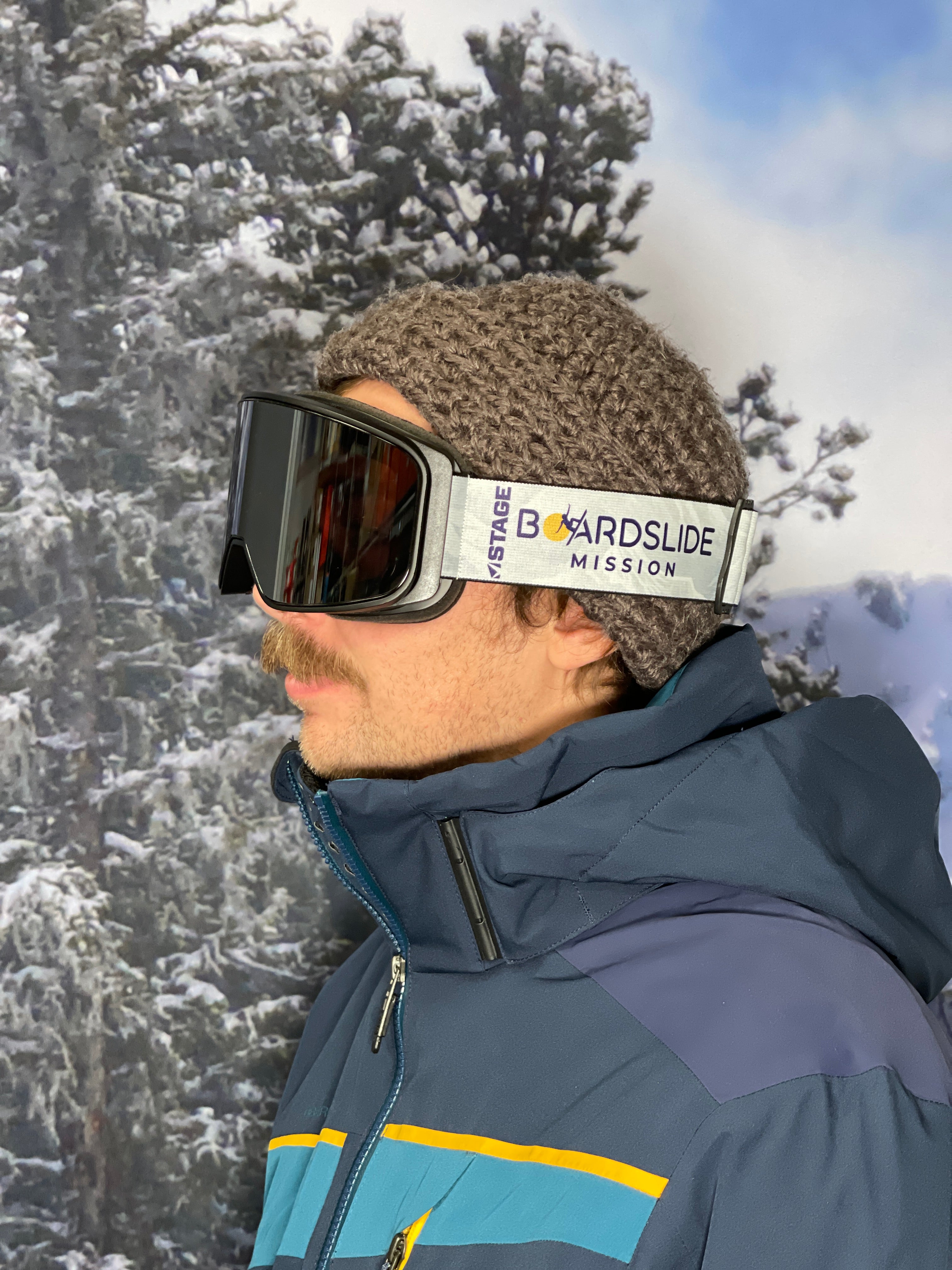 A man profile view wearing a knitted brown beanie and STAGE custom ski goggles. The goggles strap has "BOARDSLIDE MISSION" printed on it. He is dressed in a blue and gray winter jacket with yellow stripes, set against a backdrop of snow-covered trees.