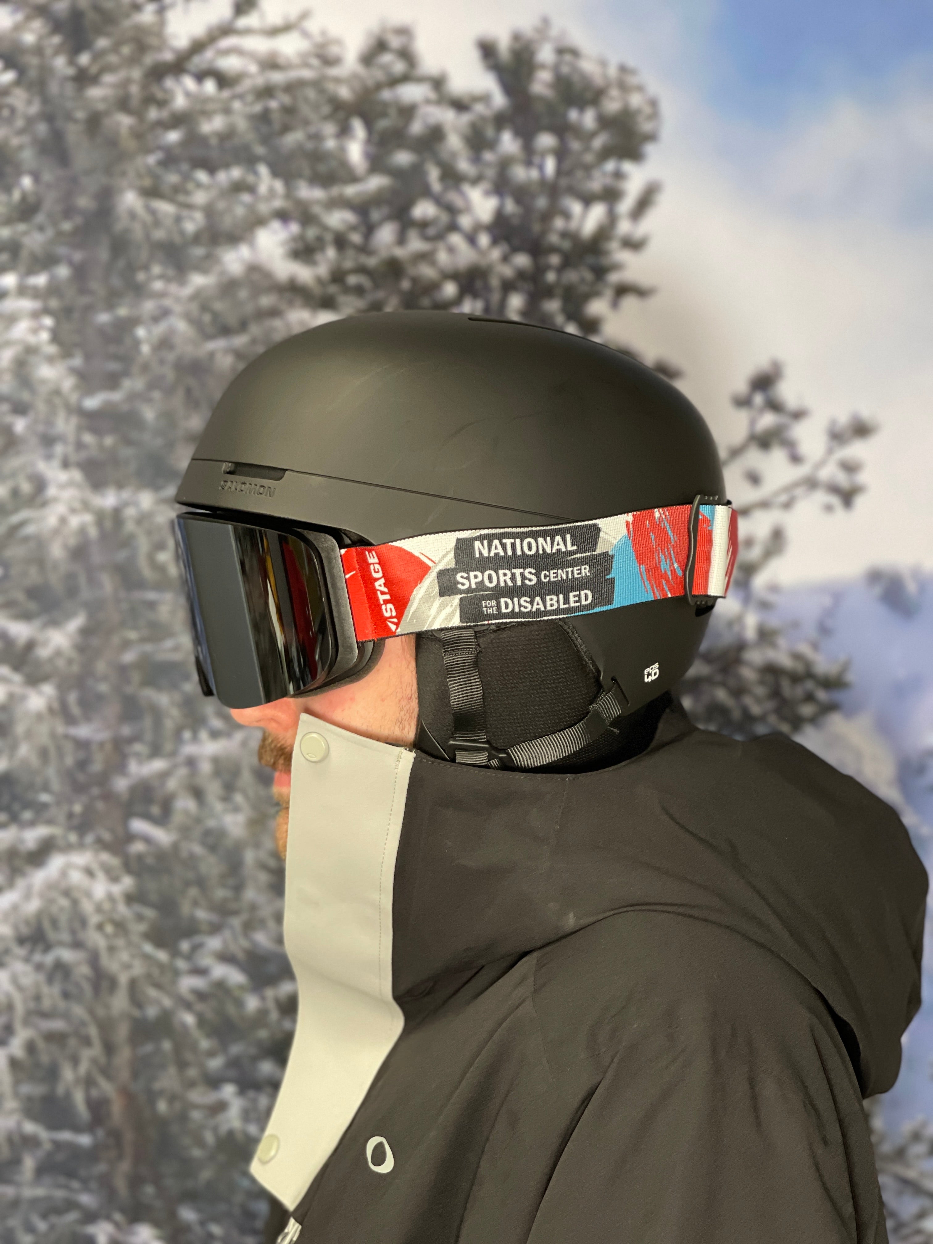A closeup of a man wearing ski gear featuring a STAGE Custom Ski Goggle showcasing the National Sports Center for the Disabled logo.