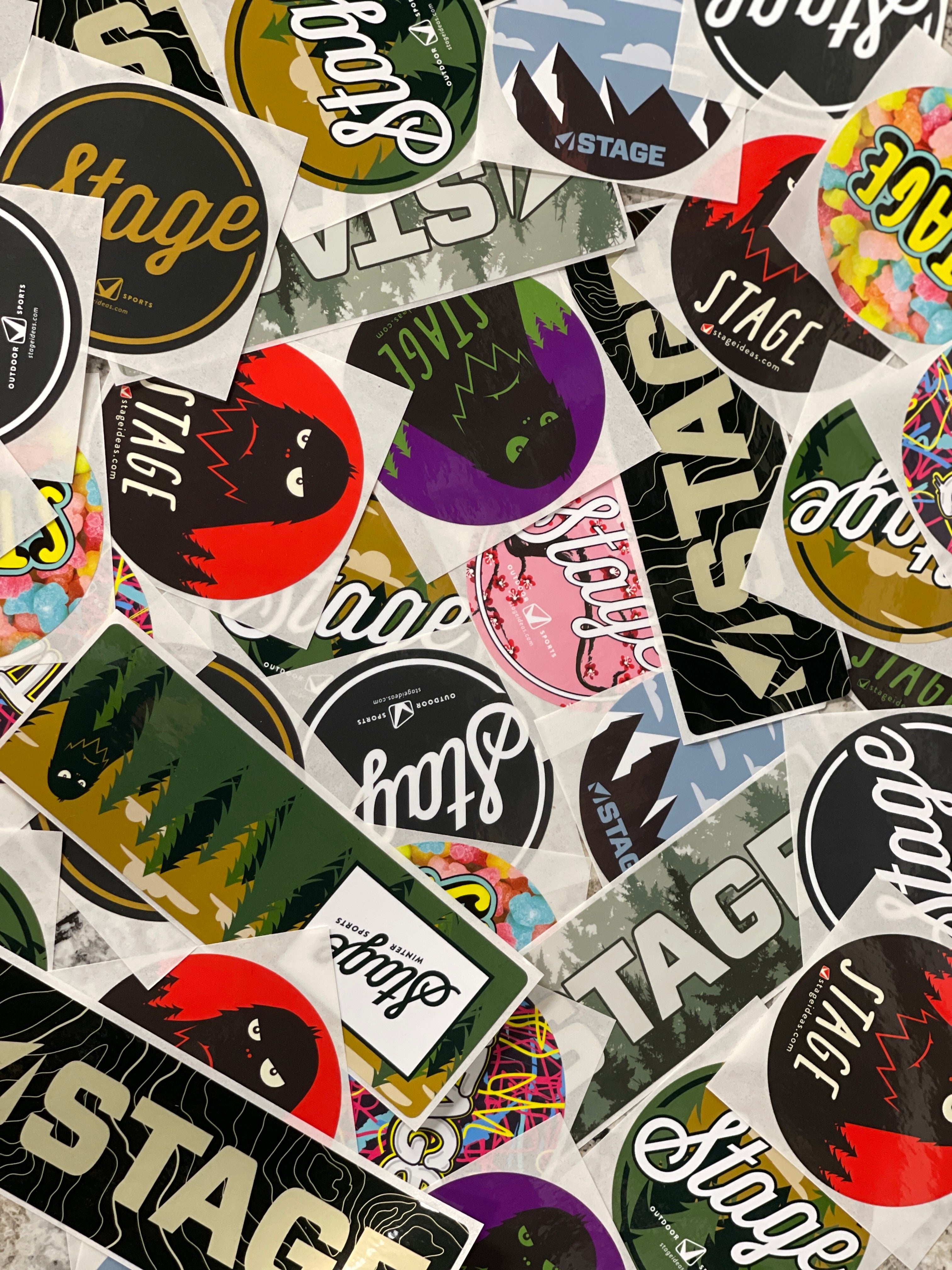 A pile of a variety of stickers featuring the STAGE Ski Goggle brand logo and promotion for custom ski goggles.