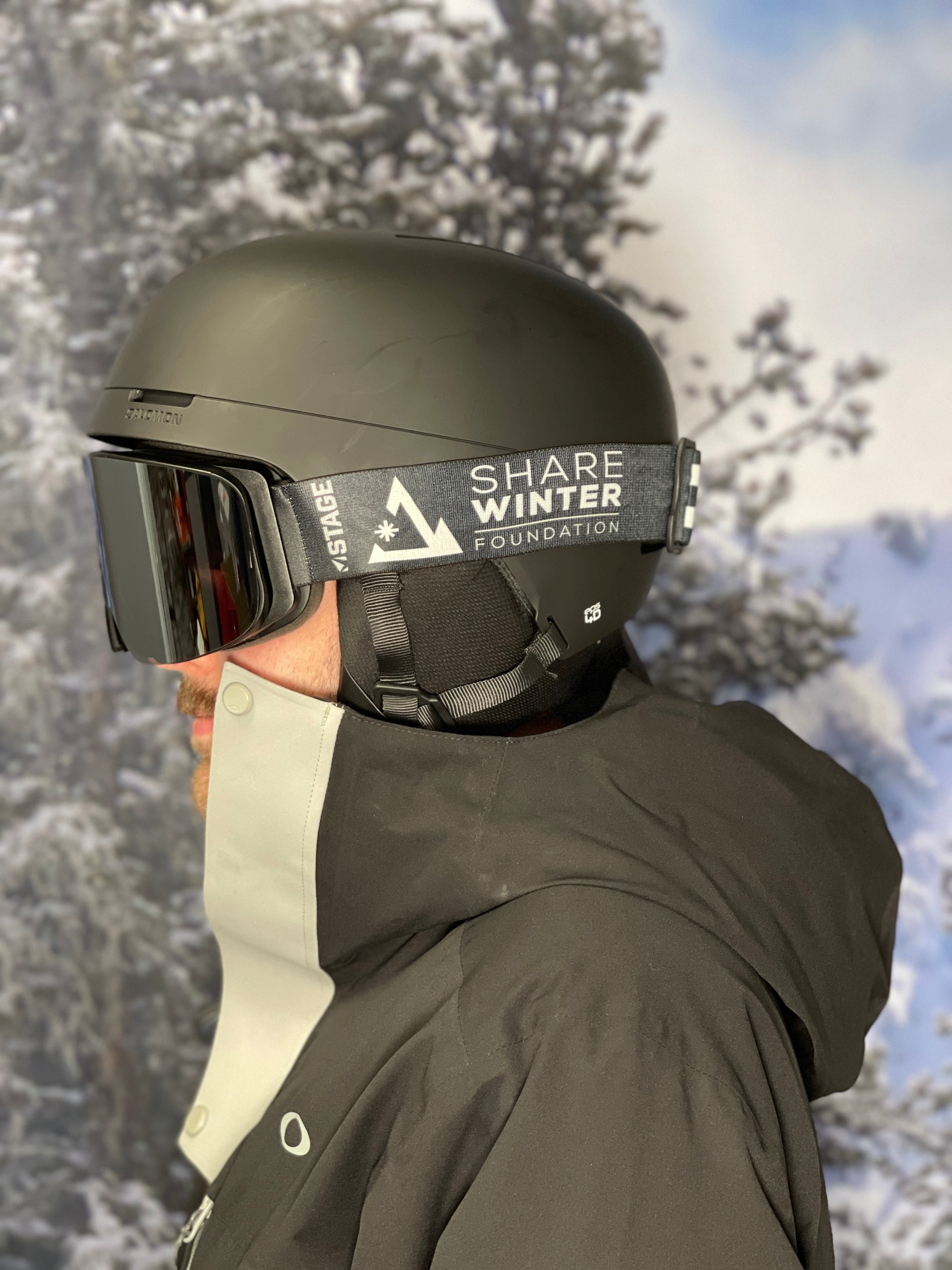 A man wearing a black ski jacket, black helmet, and STAGE Custom Ski Goggles. The snow goggles feature the Share Winter Foundation logo on the strap. The background is snowy.
