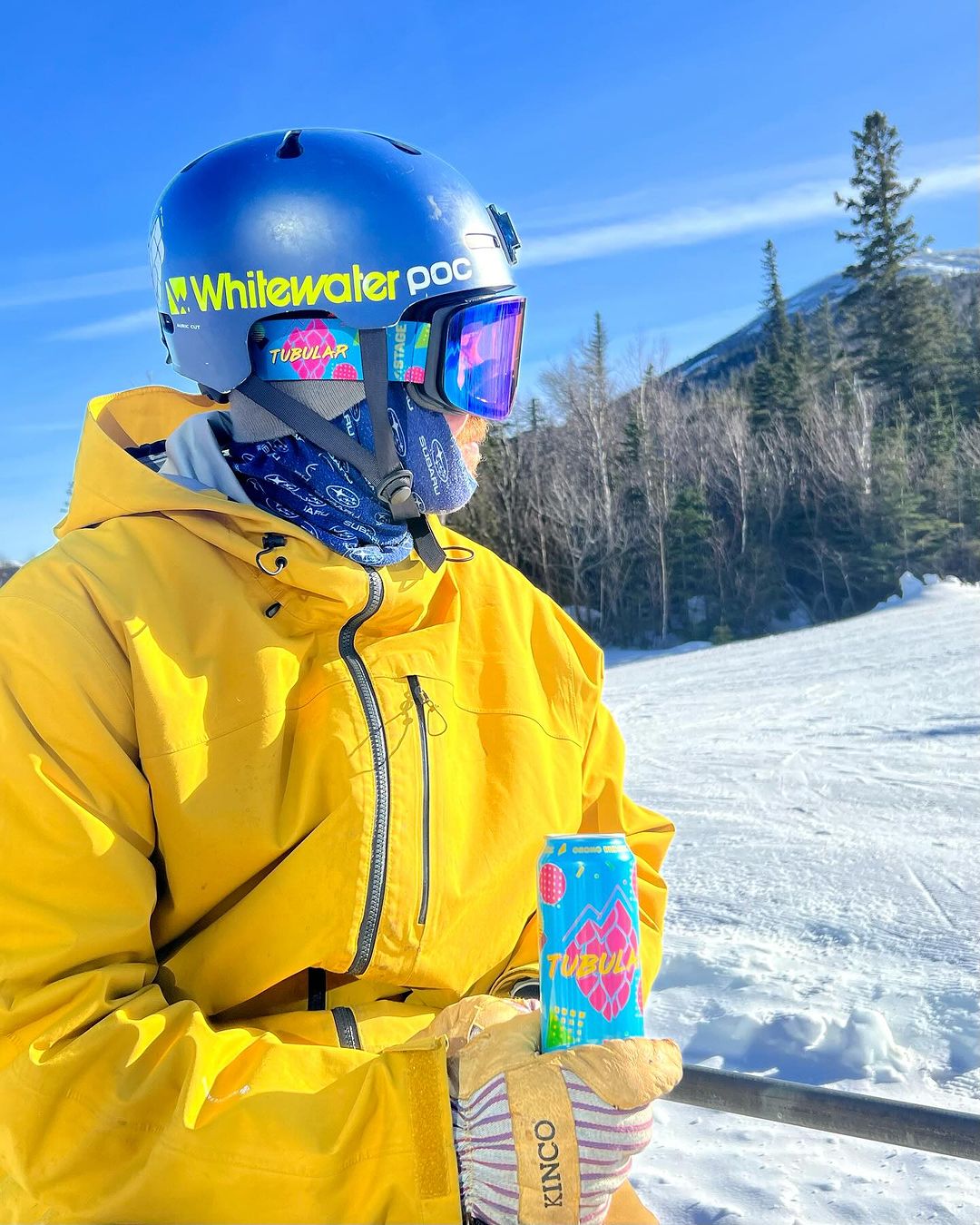 A man standing on a ski slope wearing a yellow ski jacket, blue ski helmet, and STAGE custom ski goggles featuring the Tubular IPA design from Orono Brewing