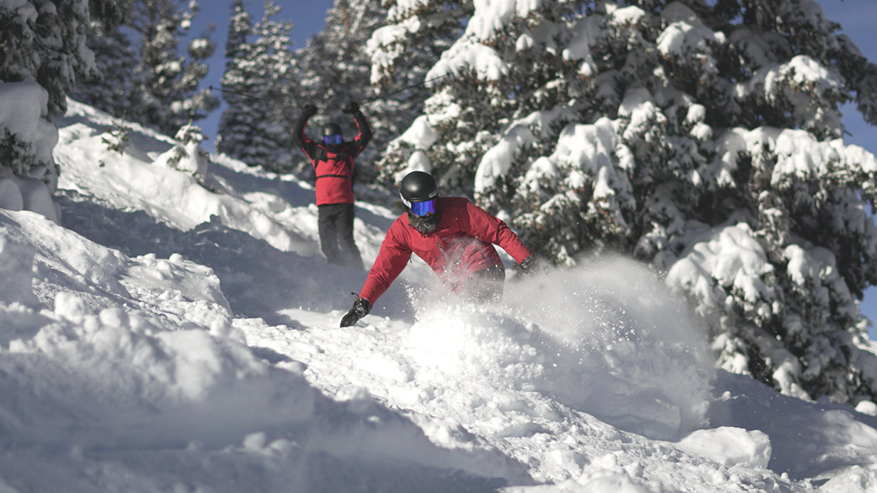 A snowboarder is snowboarding in powder while wearing STAGE's Prop Ski Goggles with Detector Lenses. Another skiier is in the background cheering the snowboarder on -- the skiier is also wearing STAGE Custom Ski Goggles.