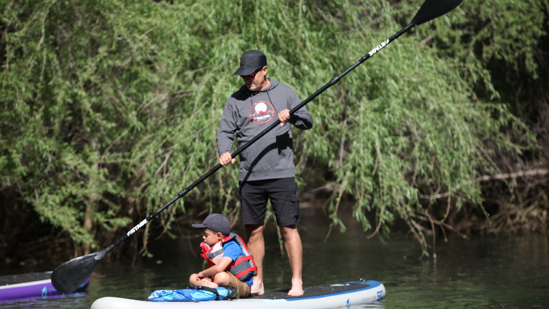 A man and a child on a STAGE Inflatable Stand-Up Paddleboard. The child is sitting on the front of the board. The man is standing on the board while paddling with the STAGE 2SIDE Double-Sided Paddleboard Paddle.