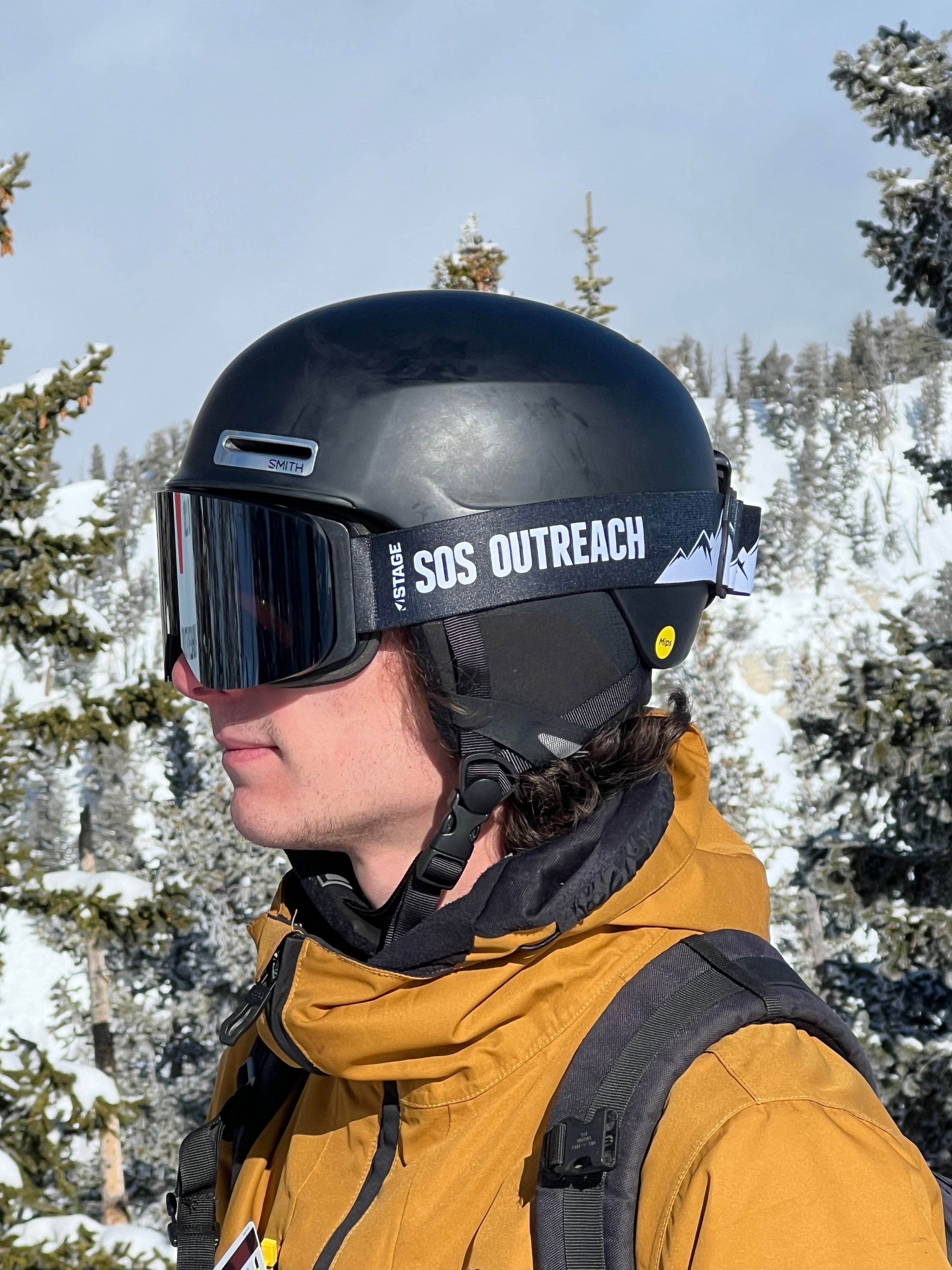 A man on the ski slopes wearing ski clothing featuring a STAGE Custom Ski Goggle showcasing SOS Outreach's logo
