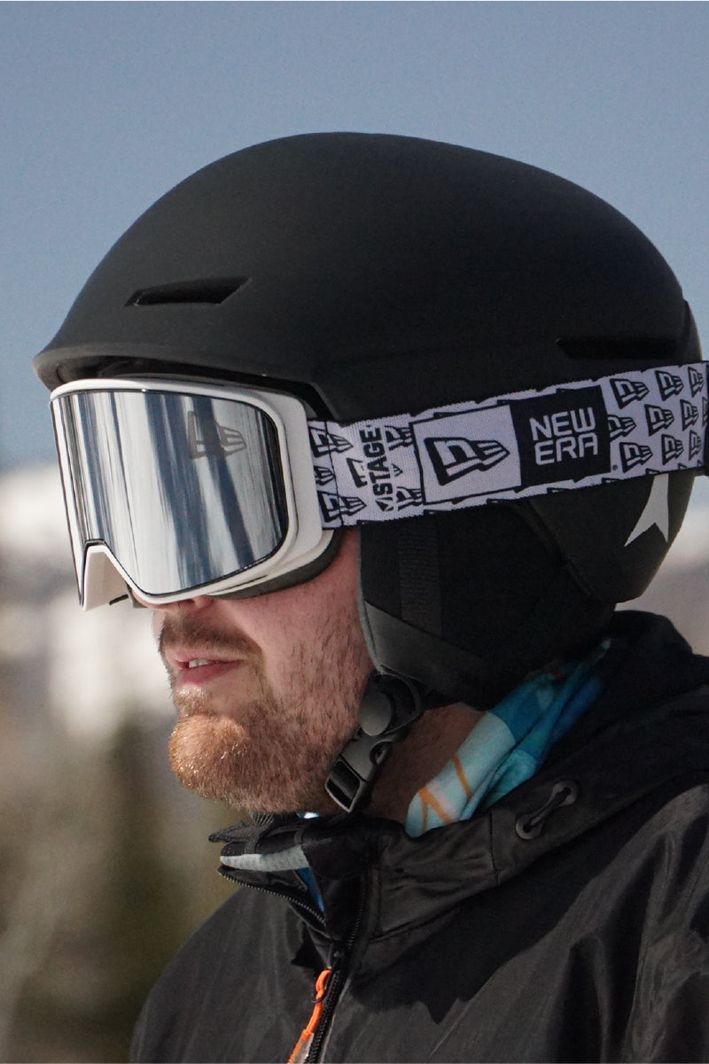 A man wearing STAGE Promo Ski goggles featuring New Era. The man is also wearing a black snowboard helmet and multi-colored face tube.