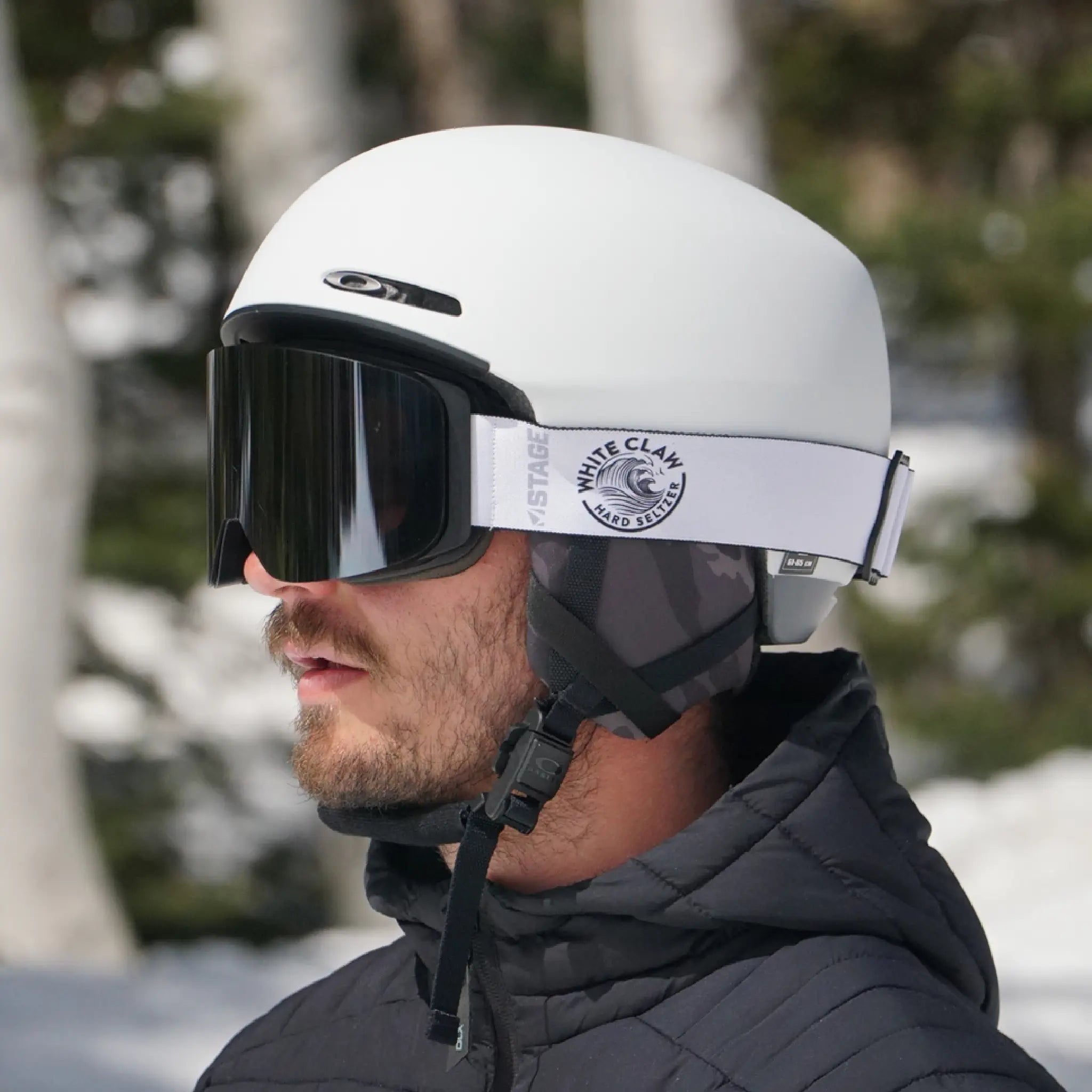 A man wearing custom ski goggles with a bold black lens and a white strap featuring the White Claw Hard Seltzer logo. He’s dressed in a black insulated jacket and white helmet with camo ear pads, standing outdoors in a snowy, wooded setting.