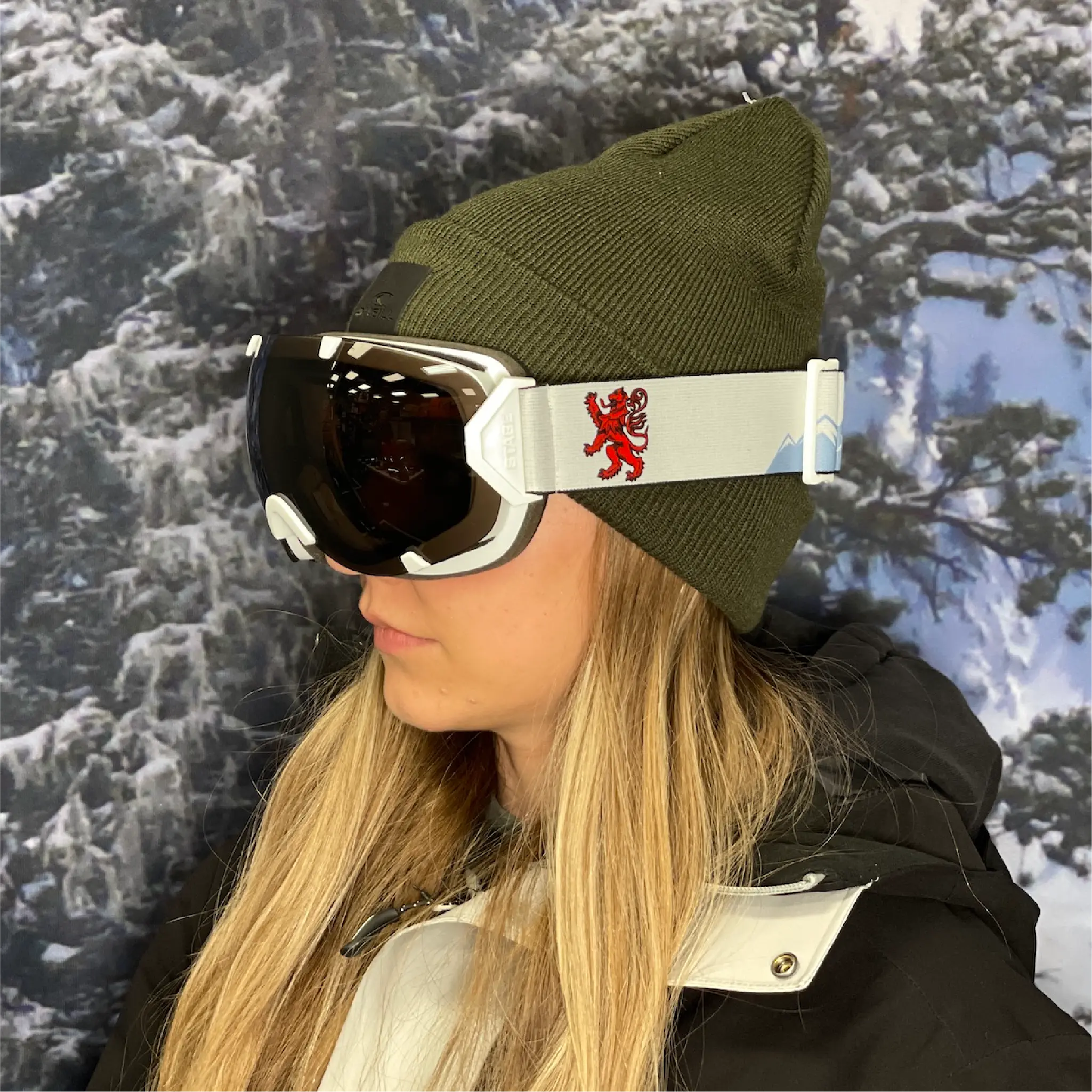 A woman wearing custom ski goggles with a white frame and a light gray strap featuring a red lion emblem and subtle mountain graphics. The dark-tinted lenses reflect indoor lighting, and she’s dressed in a black and white ski jacket with an olive green beanie, standing in front of a snowy forest backdrop.